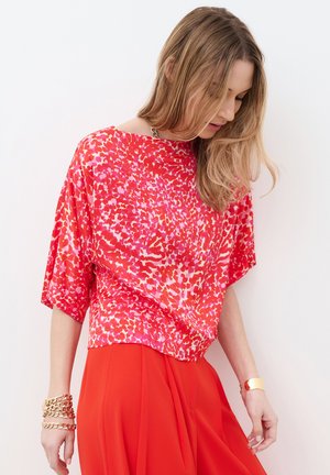 Kobieta w czerwonej bluzce w wzory i jasnoczerwonej spódnicy, patrząca w dół, z rozpuszczonym blond włosami, nosząca złote bransoletki i mankiet.