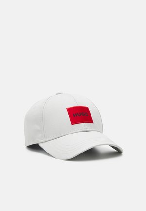 Casquette de baseball blanche avec visière incurvée et écusson rectangulaire rouge sur le devant affichant "HUGO" en lettres noires.