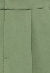 Tissu vert avec des détails de coutures verticales et une couture de poche partiellement visible sur un vêtement.