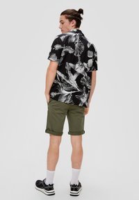 Korte mouwen zwart shirt met wit tropisch bladdessin, gecombineerd met olijfgroene opgerolde shorts en zwarte sneakers met witte accenten.