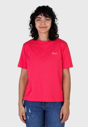 Kvinne med krøllete mørkt hår som smiler, iført en knallrosa kortermet T-skjorte og blå slitte jeans, stående foran en ensfarget bakgrunn.