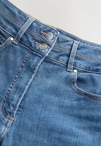 Ceinture de jean en denim bleu avec deux boutons métalliques étiquetés "NEXT DENIM", passants de ceinture visibles, poche avant et coutures détaillées.