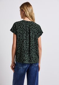 Zelený top s leopardím potiskem a krátkými rukávy, s detailem skládání na zadní straně. V kombinaci s modrými denimovými kalhotami, viděno zezadu.