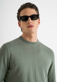 Maglione verde a maglia con scollo rotondo, abbinato a occhiali da sole neri angolari, texture liscia, design minimalista.