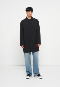 Μαύρο παλτό με ίσιο κόψιμο, ψηλό γιακά, πλαϊνές τσέπες και λείο ύφασμα, σε συνδυασμό με ανοιχτό μπλέ φαρδύ τζιν και μαύρα αθλητικά παπούτσια.