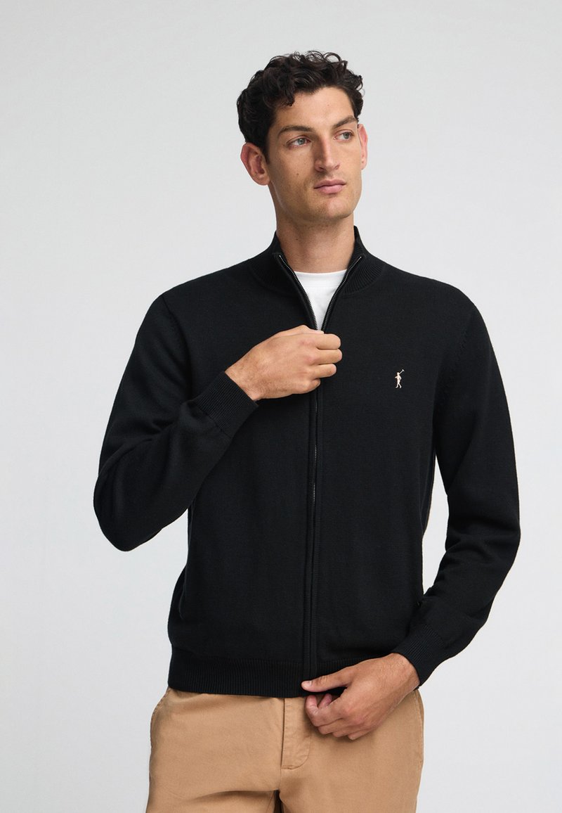 Svart zip-up kofta i stickad tyg. Har en hög krage, ribbade ärmmuddar och en liten logo på vänster bröst. Slät textur.