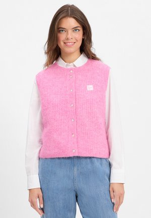 Roze gebreide vest met een ronde halslijn, knopen aan de voorkant en een klein logo label. Draagt over een witte blouse met kraag en blauwe jeans.