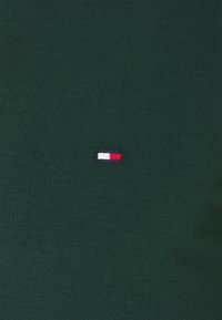 Green cotton t-shirt with a smooth texture featuring a small embroidered logo in red, white, and blue on the left chest area.