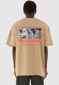 Beige bomulds t-shirt med en afslappet pasform, der har et sort-hvidt grafik af atletens ben og rød tekst, der står "BALL HARD."