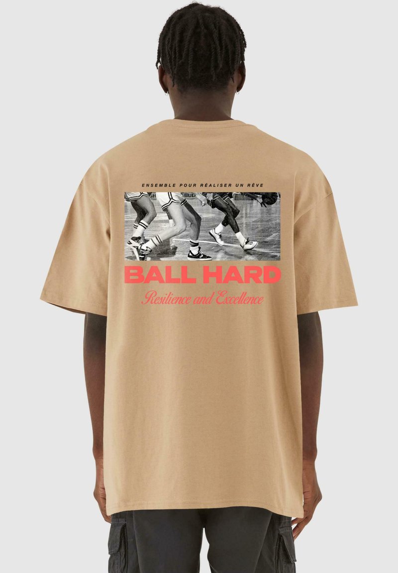 Beige bomulds t-shirt med en afslappet pasform, der har et sort-hvidt grafik af atletens ben og rød tekst, der står "BALL HARD."