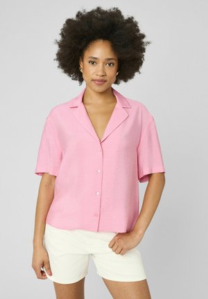 Jeune femme avec une coiffure afro naturelle portant une chemise rose ample à manches courtes boutonnée et un short blanc, debout devant un fond gris.