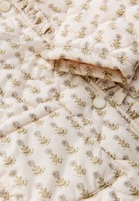Veste matelassée de couleur crème clair, arborant un motif floral en vert doux et gris. La fermeture comporte des boutons assortis et des accents froncés.