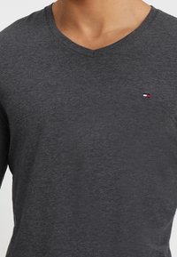 Dunkelgraues V-Ausschnitt-T-Shirt mit einem kleinen rot-weiß-blauen Flaggenlogo auf der oberen linken Brust.