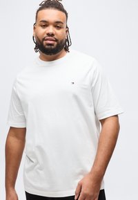 Άνδρας με γενειάδα και πλεξούδες που φοράει ένα απλό λευκό μπλουζάκι με ένα μικρό λογότυπο στο στήθος, στέκεται μπροστά σε λευκό φόντο.