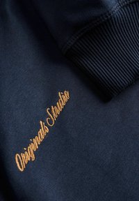 Donker marineblauwe stof met geribde manchetten; features gouden geborduurde tekst "Originals Studio." Gladde textuur met subtiele glans.