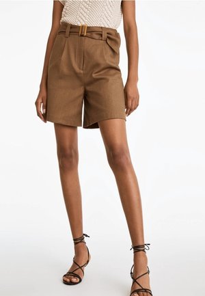 Frau trägt braune Shorts mit hohem Bund und Gürtel sowie schwarze Sandalen mit Riemchen, steht vor einem schlichten weißen Hintergrund.