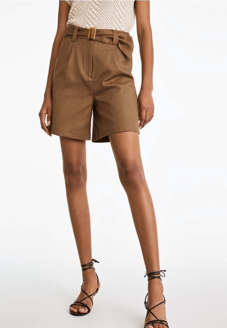 Femme portant un short taille haute marron avec ceinture et des sandales noires à brides, debout devant un fond blanc uni.