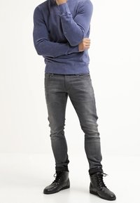 Muž stojící se založenýma rukama, oblečený v modré mikině, vybledlých šedých džínách a černých botách na šněrování na jednobarevném pozadí.