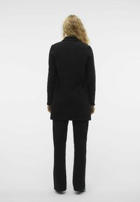 Cappotto lungo nero con vestibilità su misura, caratterizzato da una texture liscia e un design a colonna singola. Abbinato a pantaloni neri coordinati.