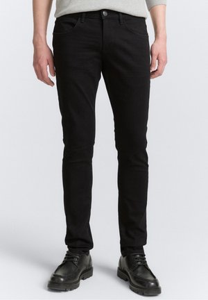 Homme portant un jean noir slim et des chaussures noires à lacets, debout devant un fond clair uni.