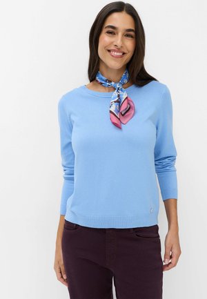 Donna che indossa un maglione azzurro chiaro, pantaloni scuri e una sciarpa blu e rosa a fantasia legata intorno al collo, sorridente verso la fotocamera.