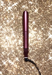 GHD krultang, cilindrisch ontwerp in metallic rozenkleur, met een zwarte kabel en merkl logo. Achtergrond van fonkelende gouden pailletten.