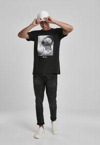 Sort t-shirt med et centralt monokrom basketballkurv grafik. Bæret med slidte mørke jeans og hvide sneakers. Hvid baseball cap holdt i hænderne.