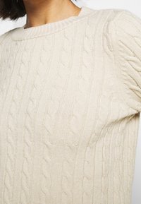 Beige Kabelstrickpullover mit Rundhalsausschnitt, der strukturierte, gewebte Muster und leicht puffierte Kurzärmel aufweist.