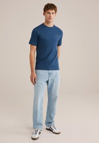 Naujai mėlynos trumpomis rankovėmis marškiniai su apvaliu kaklu ir maža logotipu, derinami su šviesiai mėlynomis tiesiomis kelnėmis ir baltais sportbačiais su juodomis juostelėmis.