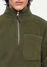Zeleni flis pulover s visokim ovratnikom, polu-zip sprijeda i džepom na prsima. Teksturalna površina i mekani završetak.