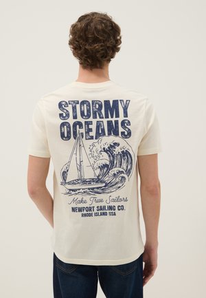 Uomo che indossa una maglietta bianca con stampa blu navy di una barca a vela, grandi onde oceaniche e testo "Stormy Oceans, Make True Sailors, Newport Sailing Co."