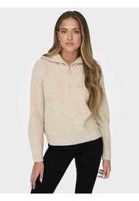 Pull en maille beige avec un col zippé mi-haut, texture côtelée et manches longues. Présente un ourlet large et un anneau en métal argenté pour la tirette du zip.