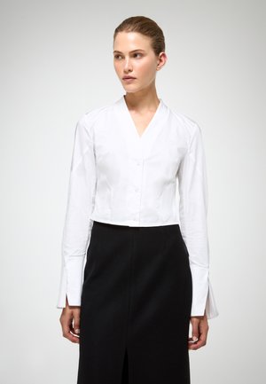 Camicia bianca cropped con colletto a V, maniche lunghe e dettagli testurizzati ai polsini, abbinata a una gonna nera a vita alta con uno spacco.