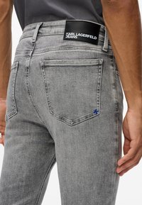 Pelēki džinsi ar šauru piegriezumu, ar melnu ādas etiķeti un mazu zilu akcentu aizmugurējā kabatā. Klasisks 5 kabatu dizains.