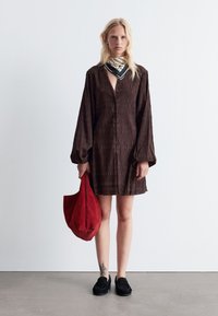 Braunes, strukturiertes Kleid mit Knopfverschluss und Ballonärmeln; kombiniert mit einem schwarzen gemusterten Schal und einer roten Wildledertasche. Schwarze Slipper.