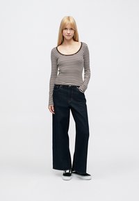 Csíkos, hosszú ujjú ing bézs és barna színekben, széles szárú sötét farmernadrággal kombinálva. A modell az egyik kezét a zsebében tartja, fekete tornacipőt visel.