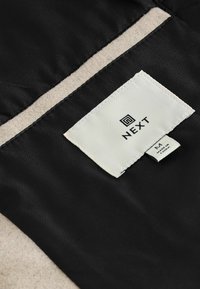 Czarna tkanina o gładkiej strukturze, z kremową metką na której widnieje napis "NEXT" oraz rozmiar "M" w czarnym druku. Wykończona beżowym obszyciem.