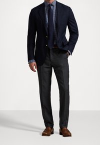 Marinblå blazer med guldknappar, rutig blå skjorta, mörkgrå byxor och bruna läderskor med loafermodell.