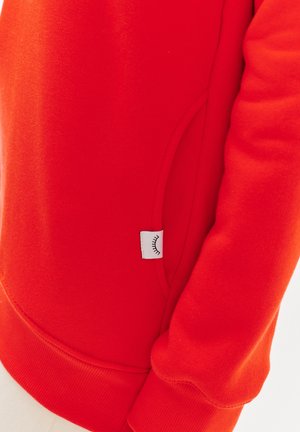 Červená mikina s měkkou texturou, vybavená boční kapsou a šitým lemem. Malá bílá visačka s grafikou oka po straně.