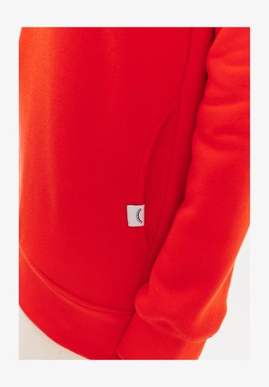 Czerwony sweter z miękką fakturą, z boczną kieszenią i przyszytym wykończeniem. Mała biała metka z graficznym motywem oka po jednej stronie.