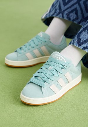 Jasnoniebieskie tenisówki Adidas z białymi paskami i gumową podeszwą, noszone z białymi skarpetkami i niebieskimi spodniami w wzory na zielonej powierzchni.