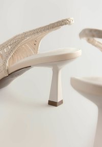 Beige sandaalit korokkeella, joissa on punottu päällinen, avonainen varvas, kaareva muotoilu ja hoikka, kulmikas korko ruskealla yksityiskohdalla pohjassa.