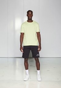 Šviesiai žalia trumpomis rankovėmis palaidinė su siuvinėtu "Champion" logotipu, derinama su juodais šortais ir baltais sportbačiais, stovint ant veidrodinio paviršiaus.