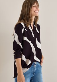 Blusa viola scuro con motivo astratto bianco, maniche lunghe e vestibilità comoda. Presenta una scollatura a V e polsini arrotolati. Abbinata a jeans blu.