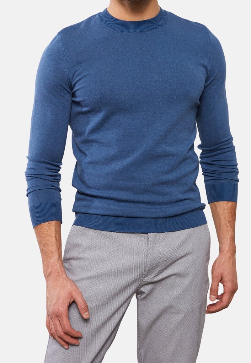 Blå stickad tröja med rund hals och ribbade muddar. Figursydd design, tillverkad av mjukt material, bärs över ljusgrå byxor.