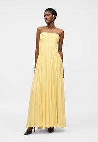 Žena v žlutých dlouhých šatech bez ramínek s řasením, doplněných černými špičatými vysokými podpatky, stojící před jednobarevným bílým pozadím.