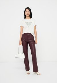 Camiseta blanca de manga corta con un patrón triangular texturizado, combinada con pantalones de cuero morados. Sosteniendo un pequeño bolso blanco.