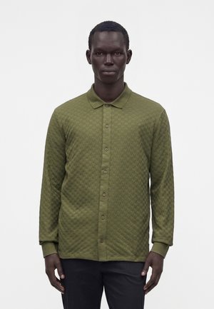 Homme portant une chemise texturée vert olive à manches longues, boutonnée et avec un col, debout devant un fond clair uni.