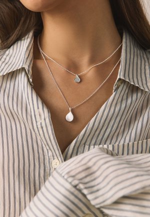 Woman wearing layered silver necklaces with teardrop pendants over a striped button-up shirt collar.