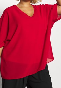 Blouse rouge légère avec une coupe ample, col en V et manches courtes. Texture lisse avec un léger éclat, design superposé à l'ourlet.